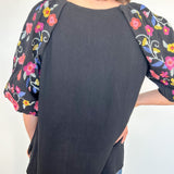 Black baseball-style shirt with floral sleeves worn by a person against a white wall.