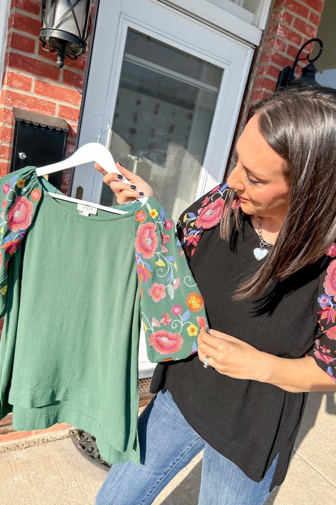 Woman holding a green top with floral sleeves in front of a brick building.