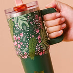 Hand holding a green floral-patterned tumbler with a pink lid against a beige background
