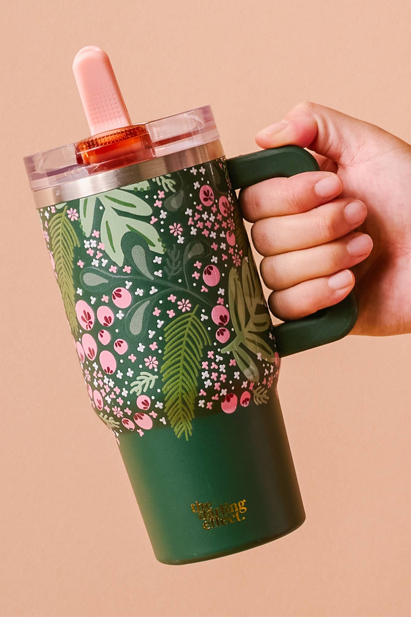 Hand holding a green floral-patterned tumbler with a pink lid against a beige background