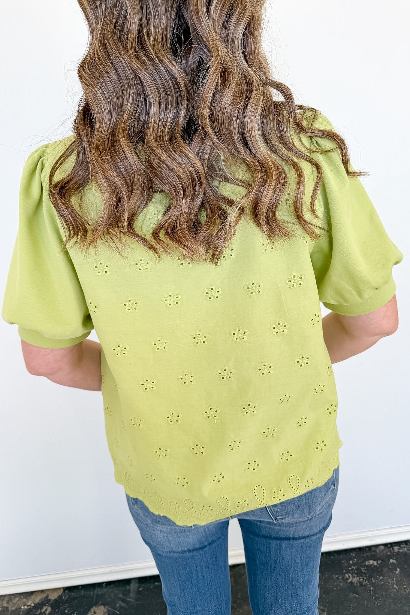 Person wearing a light green blouse with floral eyelet design and blue jeans against a white background