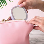 Person placing a compact mirror into a pink makeup bag on a marble surface with a plant in the background.