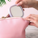 Person placing a compact mirror into a pink makeup bag on a marble surface with a plant in the background.