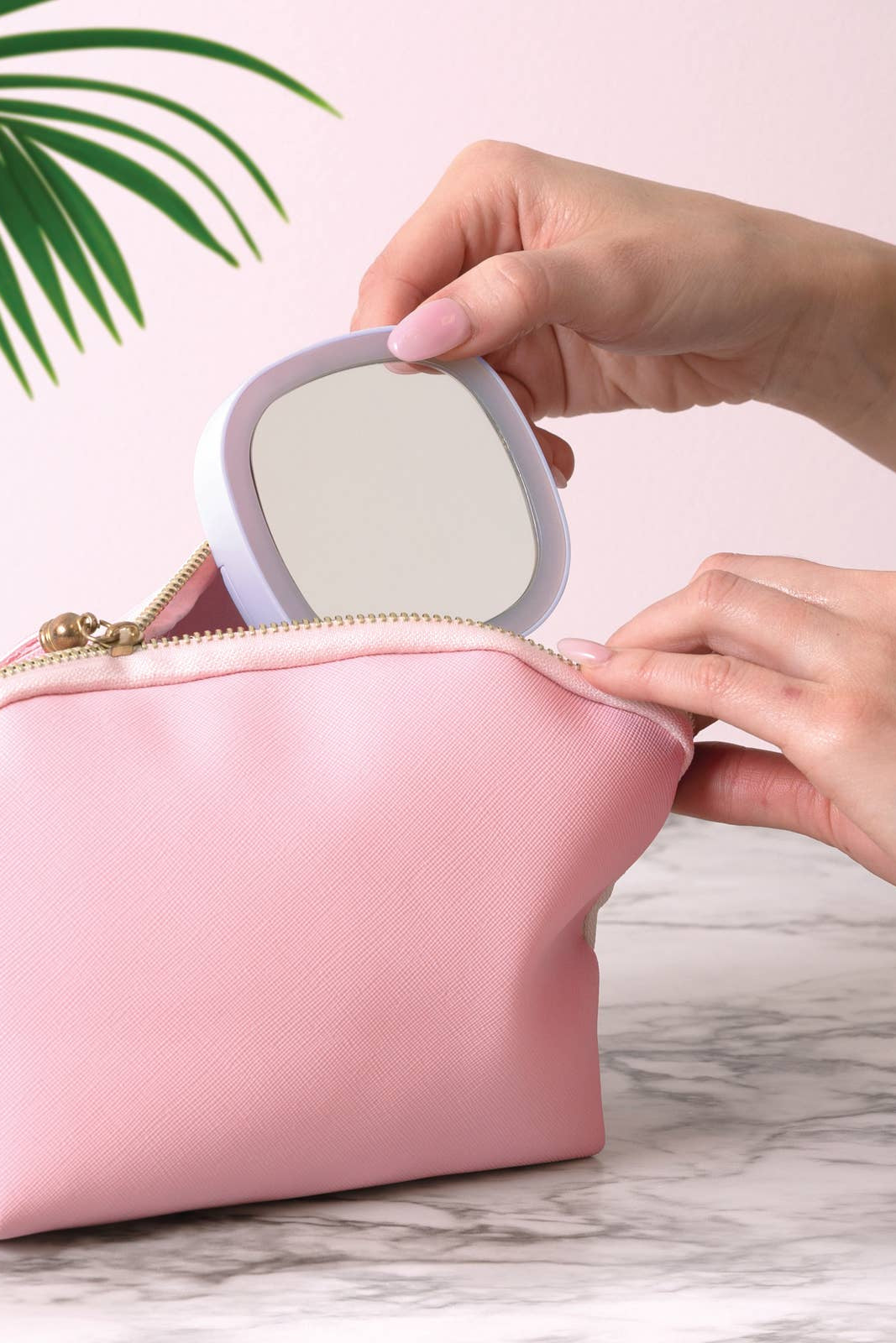 Person placing a compact mirror into a pink makeup bag on a marble surface with a plant in the background.