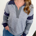 Woman wearing a gray and navy long-sleeve top with blue pants against a white wall.