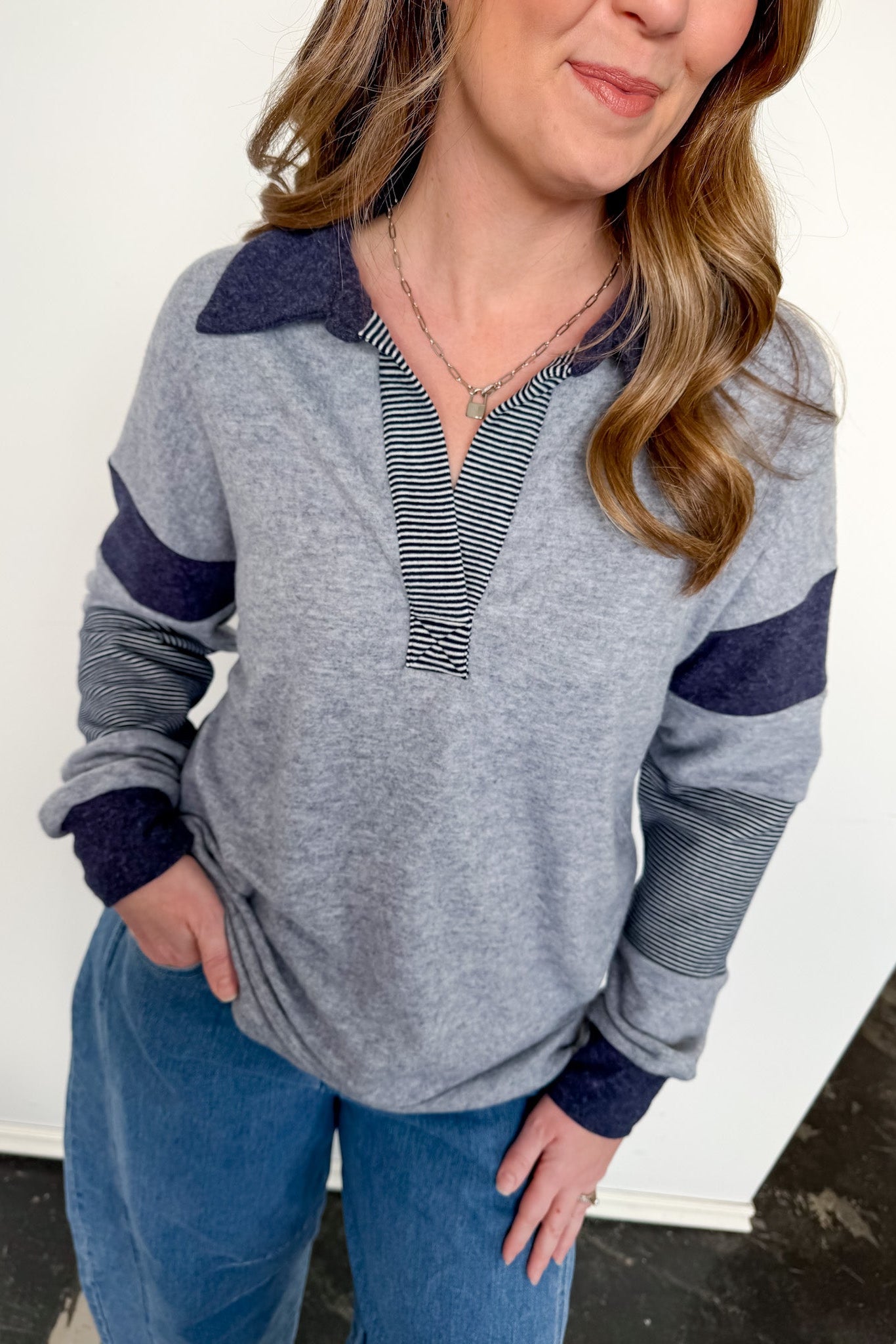 Woman wearing a gray and navy long-sleeve top with blue pants against a white wall.