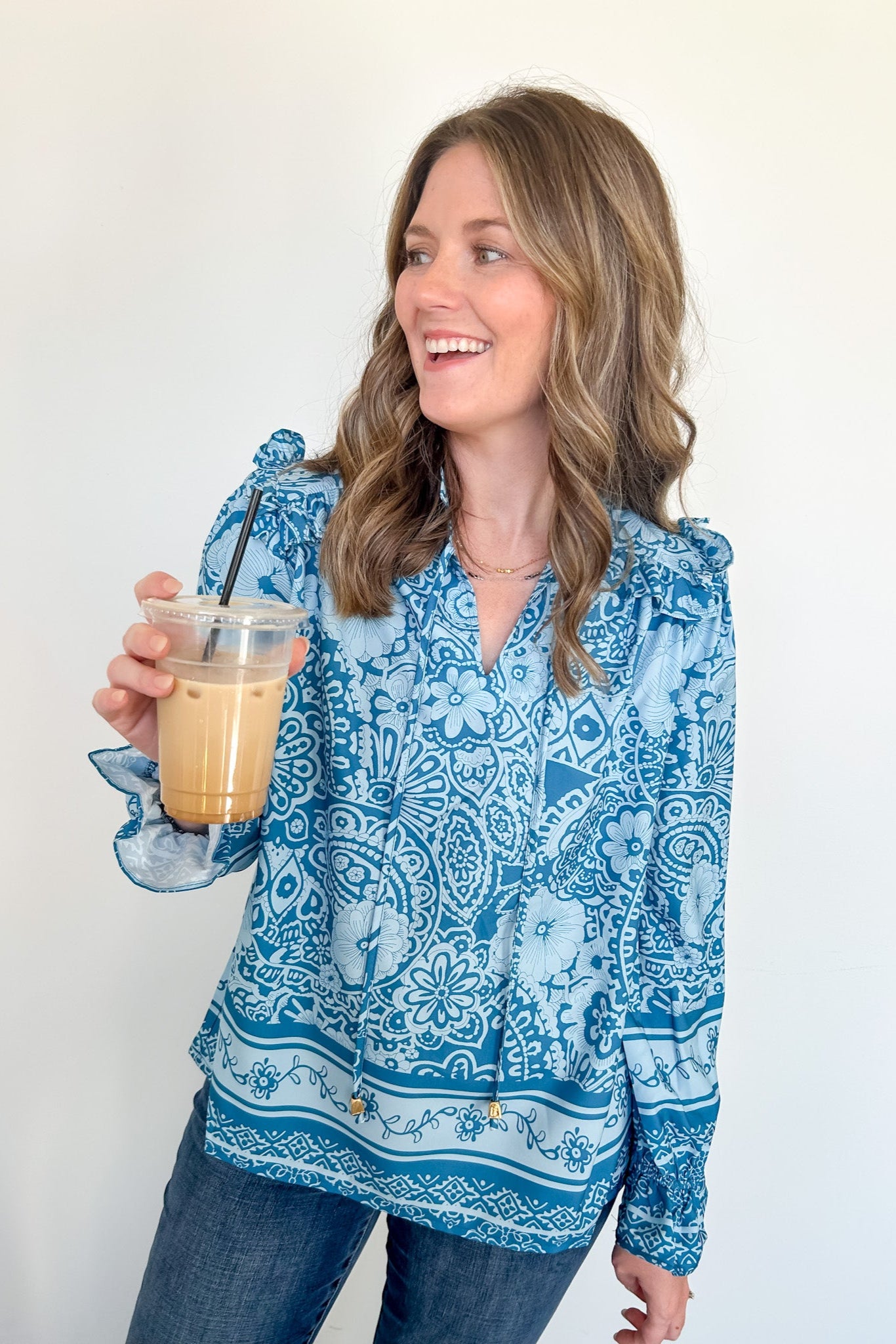 Woman wearing a blue patterned blouse holding a glass of iced coffee against a white background