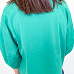 Person wearing a green sweater and blue jeans against a white wall.