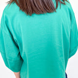 Person wearing a green sweater and blue jeans against a white wall.
