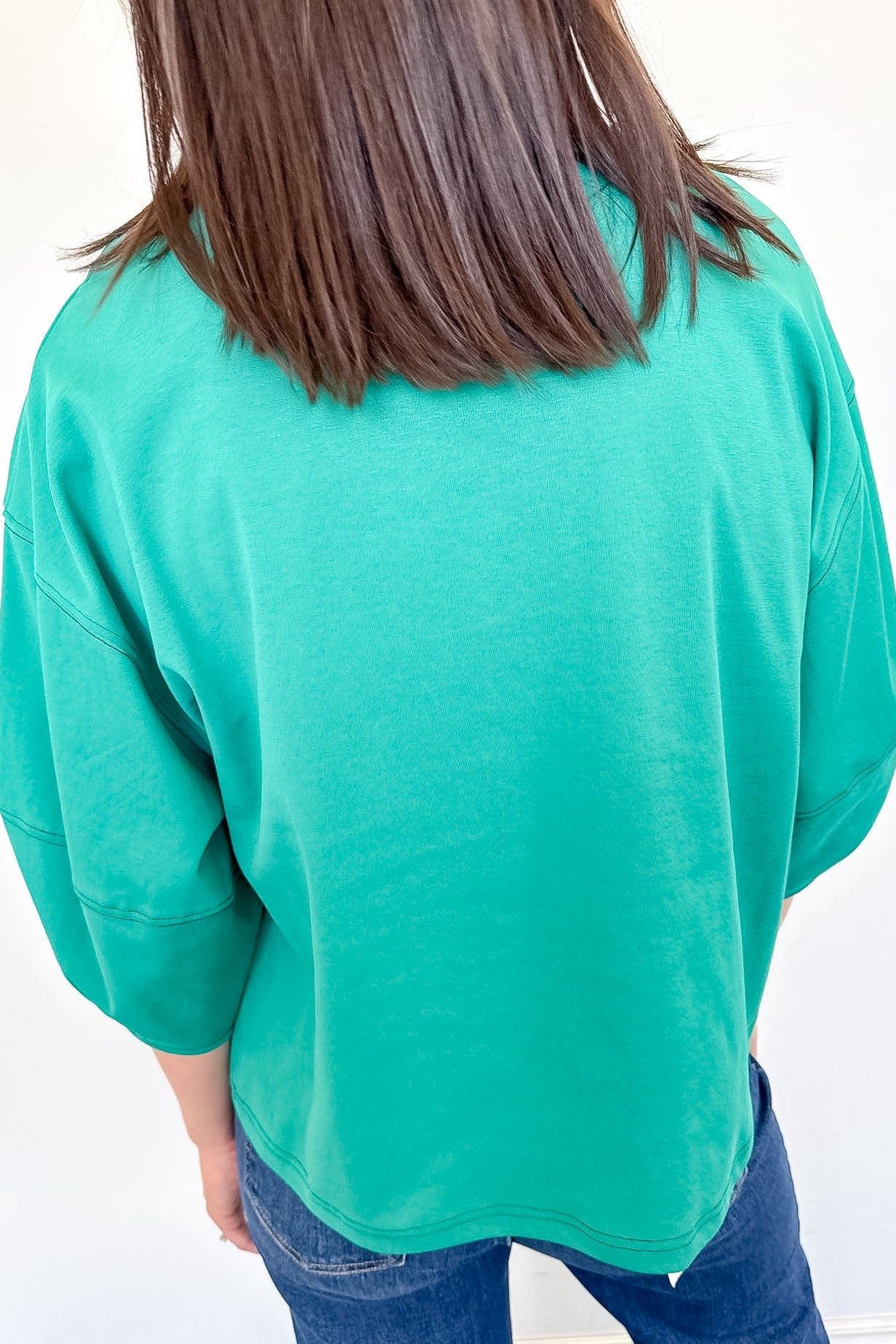 Person wearing a green sweater and blue jeans against a white wall.