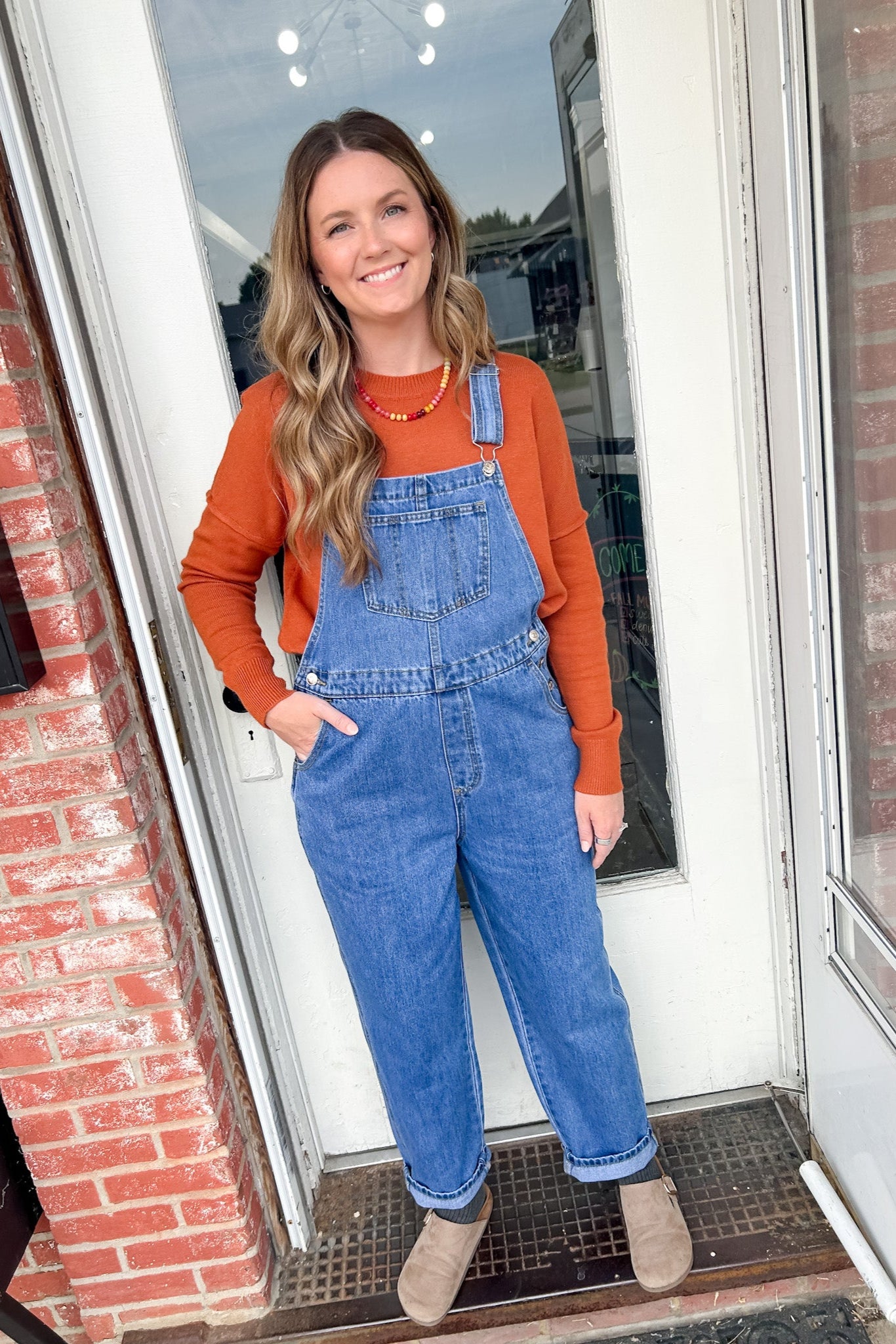 Woman wearing blue overalls and an orange shirt standing in front of a door.