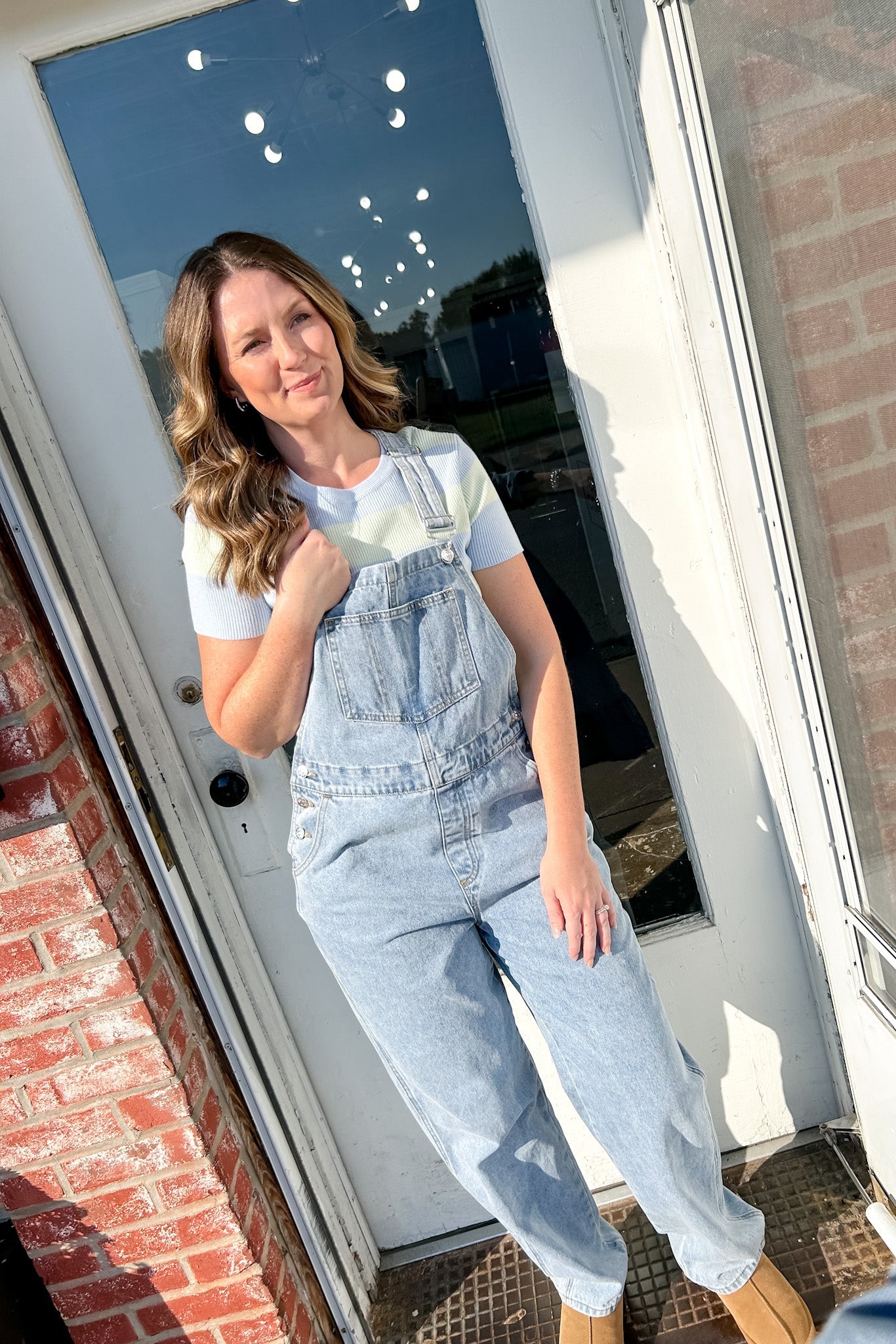 Woman wearing light blue overalls standing in front of a door with a brick wall background