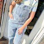 Person wearing light blue denim overalls standing on a porch.
