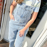 Person wearing light blue denim overalls standing on a porch.