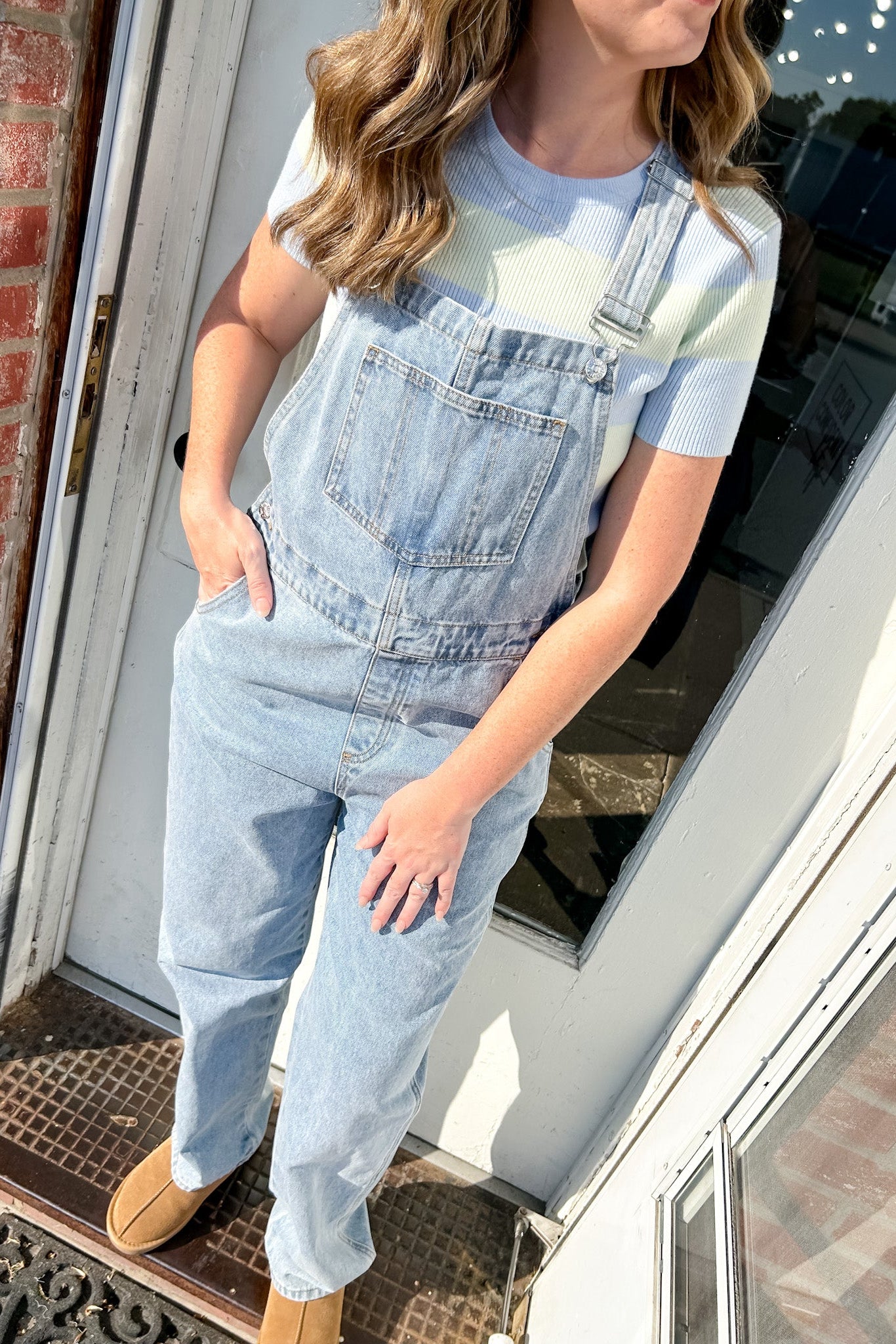 Person wearing light blue denim overalls standing on a porch.