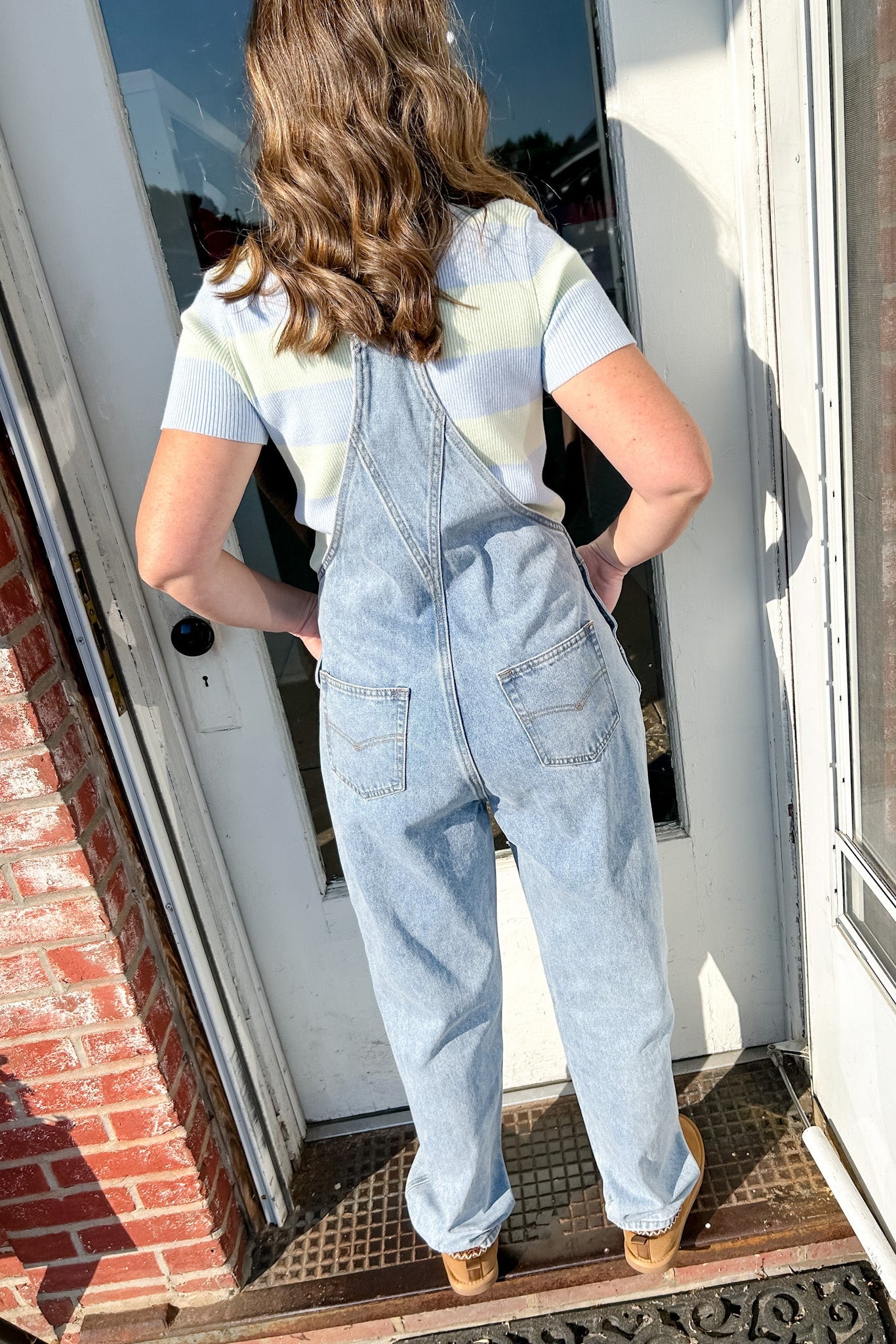 Person wearing light blue denim overalls standing on a porch.