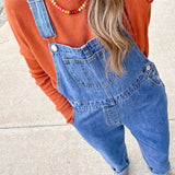 Person wearing an orange sweater and blue denim overalls on a sidewalk.