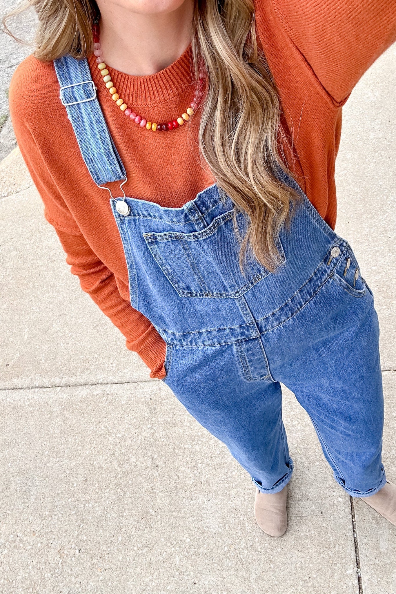 Person wearing an orange sweater and blue denim overalls on a sidewalk.