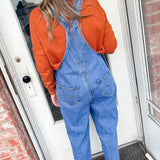 Person wearing blue denim overalls and an orange shirt standing in front of a door.