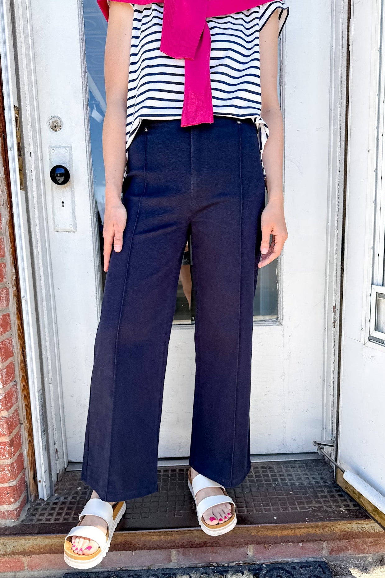 Person wearing a striped shirt with a pink bow, navy pants, and white sandals standing in front of a door.