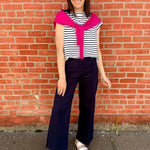 Woman wearing a striped shirt with a bright pink collar and navy pants standing against a brick wall.