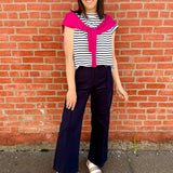 Woman wearing a striped shirt with a bright pink collar and navy pants standing against a brick wall.