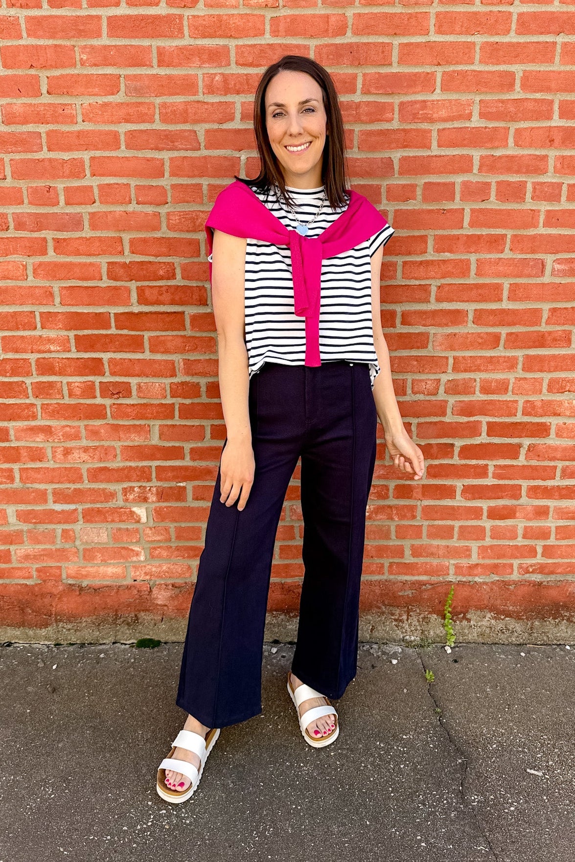Woman wearing a striped shirt with a bright pink collar and navy pants standing against a brick wall.