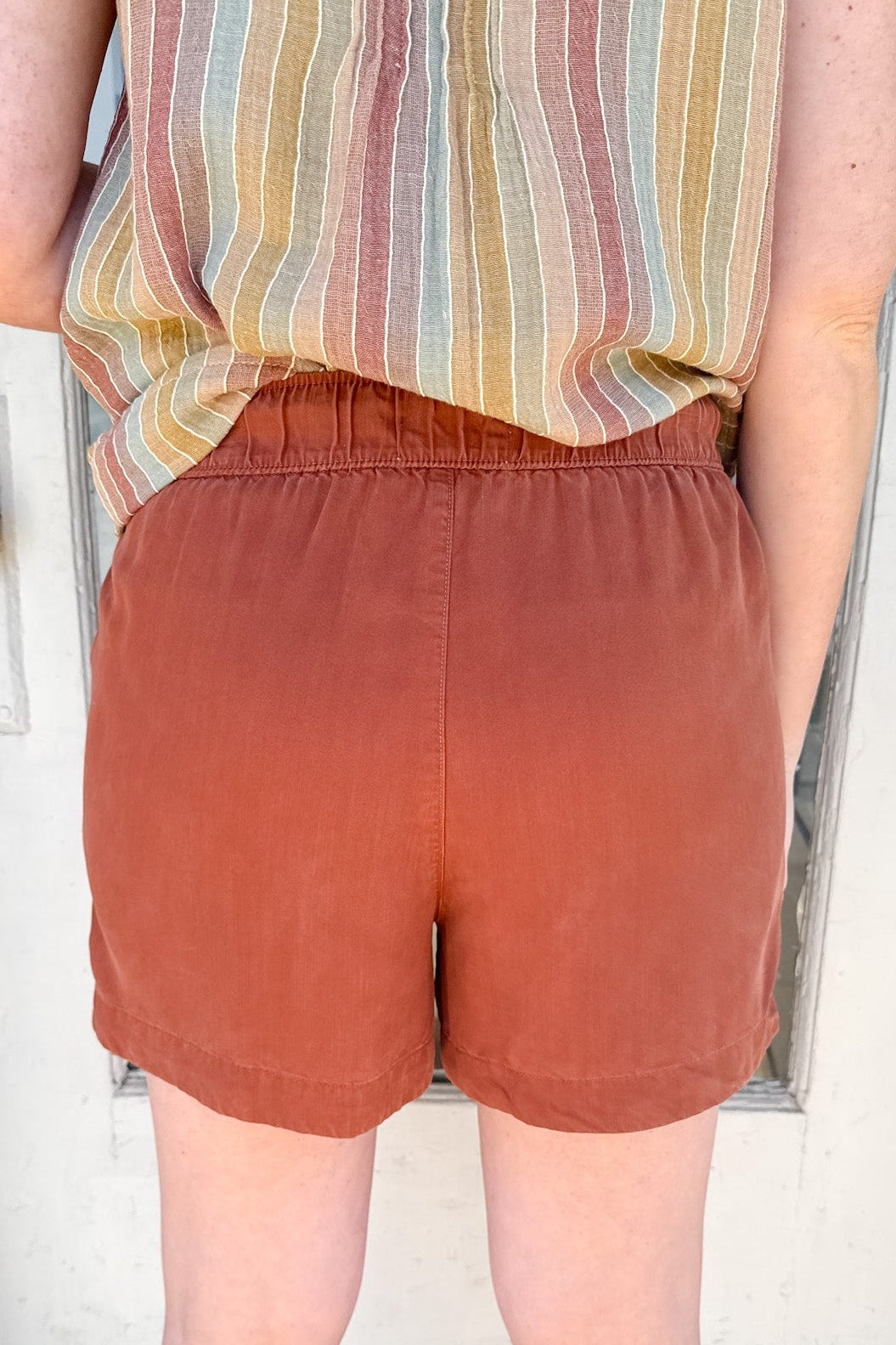 Person wearing rust-colored shorts and a striped shirt standing in front of a white door.