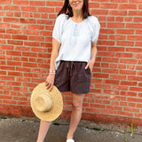 Woman standing in front of a red brick wall with a white shirt and black shorts with a straw hat in her hand.