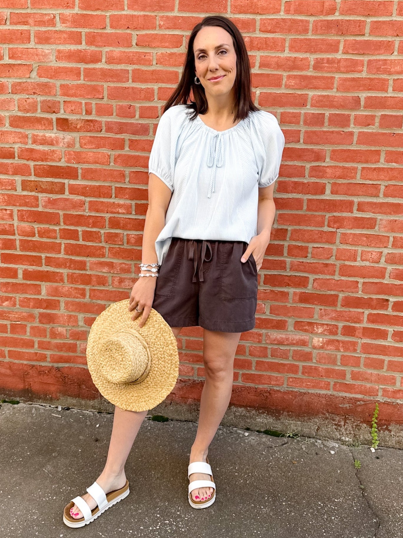 Woman standing in front of a red brick wall with a white shirt and black shorts with a straw hat in her hand.