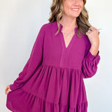 Woman wearing a short purple dress with long sleeves against a white wall.