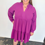 Dark haired woman wearing a purple long sleeve dress with nude heels against a white wall.
