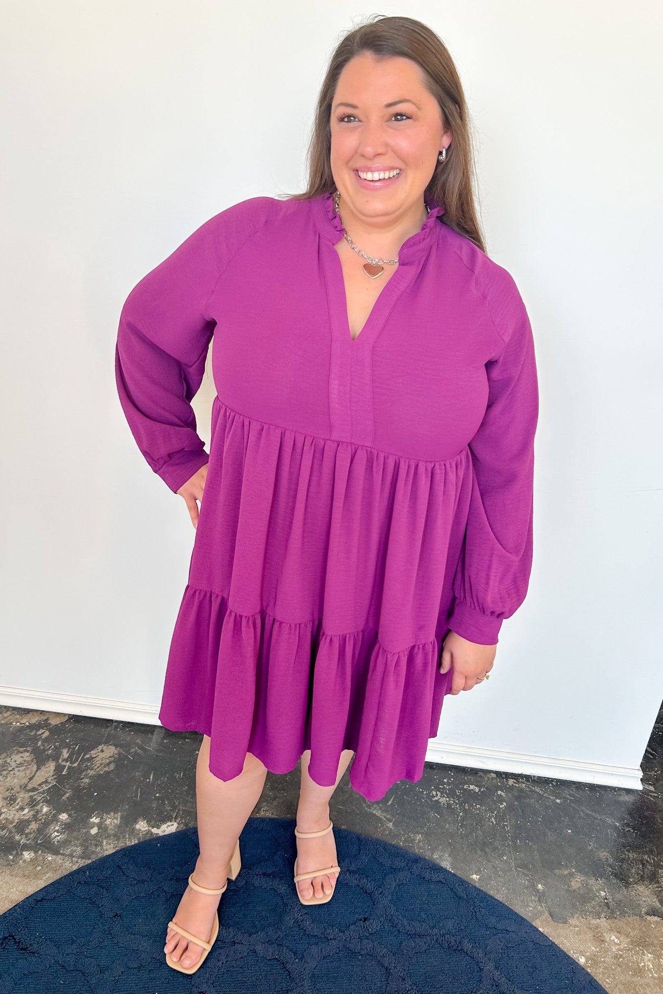 Dark haired woman wearing a purple long sleeve dress with nude heels against a white wall.