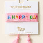 Pink woven word bracelet with 'HAPPY DAY' text on a white card with brand name, on a beige background.