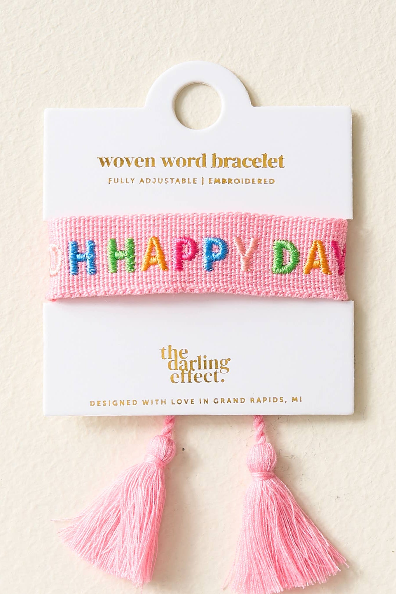 Pink woven word bracelet with 'HAPPY DAY' text on a white card with brand name, on a beige background.