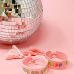 Pink bracelets with colorful text next to a disco ball on a pink background