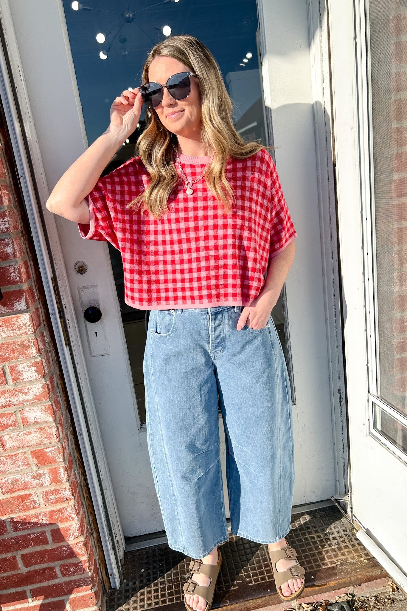 Woman wearing a red checkered shirt and blue jeans standing in front of a door.