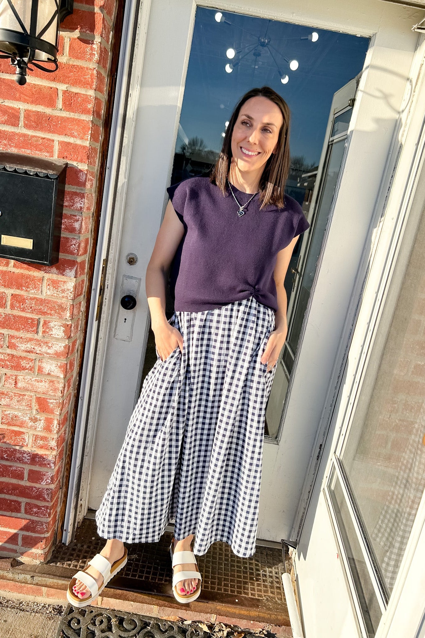 Woman standing outside a house wearing a purple top and checkered skirt.