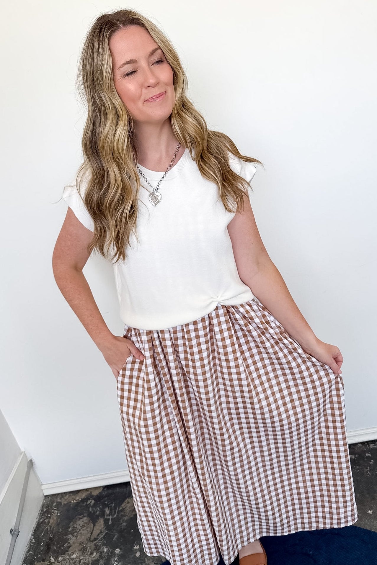 Woman wearing a white top and brown checkered skirt against a white wall.