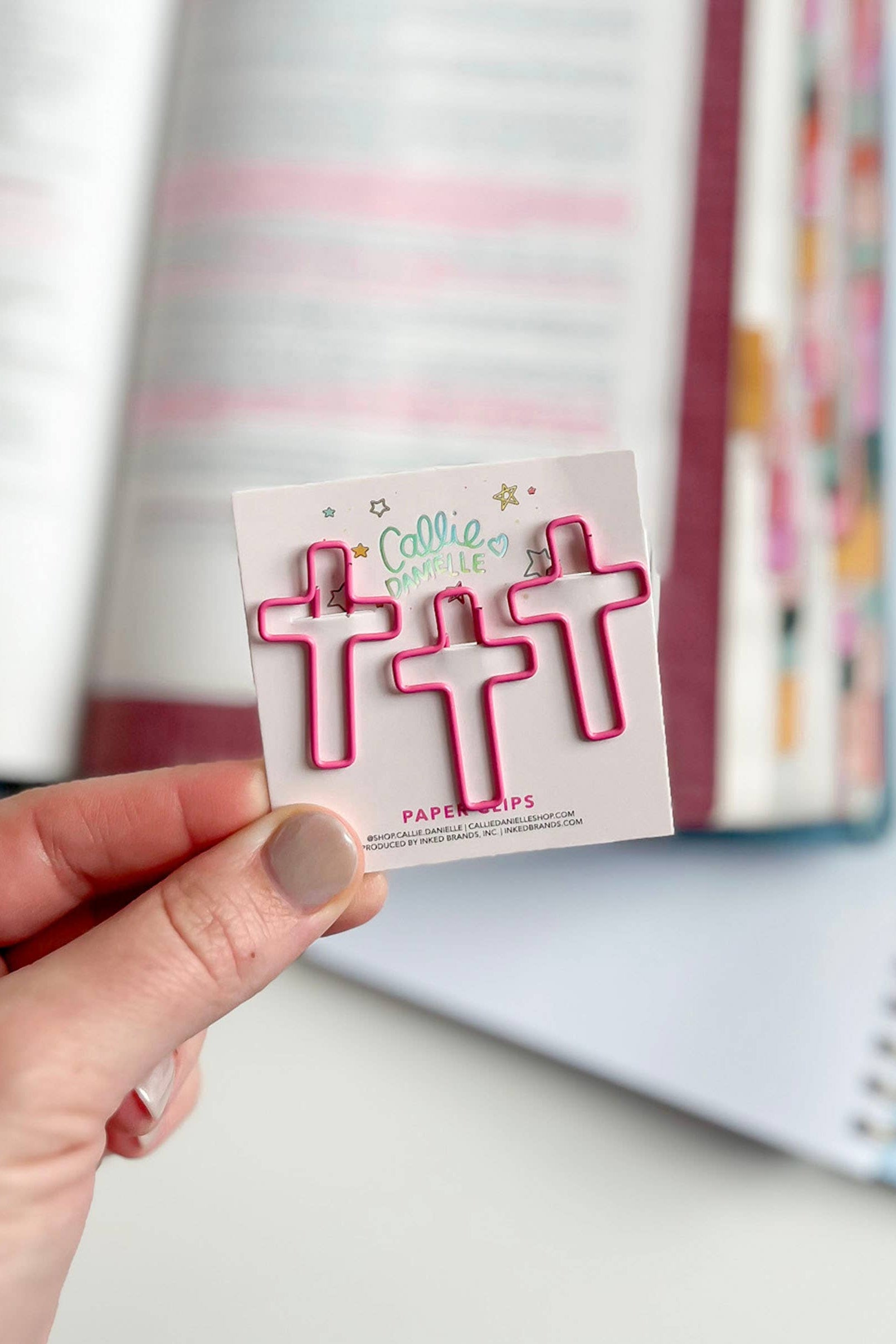 Hand holding a pack of pink paper clips with a branded label in front of a colorful notebook.