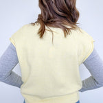 Person wearing a light yellow sleeveless sweater over a gray long-sleeve shirt with blue jeans on a white background