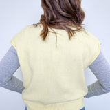 Person wearing a light yellow sleeveless sweater over a gray long-sleeve shirt with blue jeans on a white background