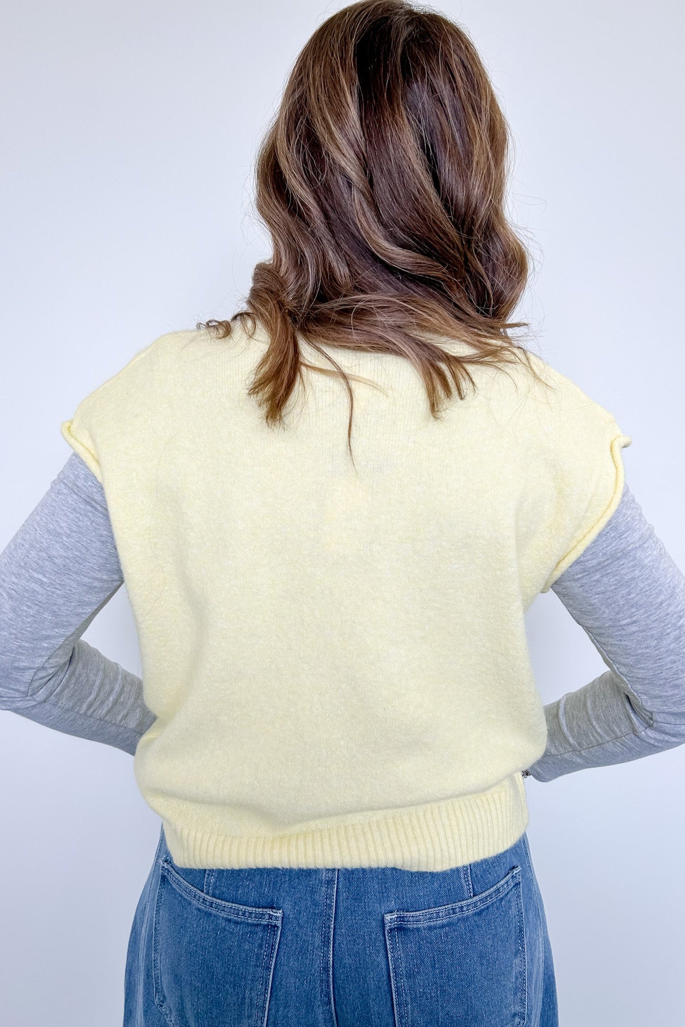 Person wearing a light yellow sleeveless sweater over a gray long-sleeve shirt with blue jeans on a white background