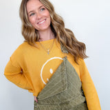 Woman wearing a yellow sweater with a smiley face and green overalls against a white background
