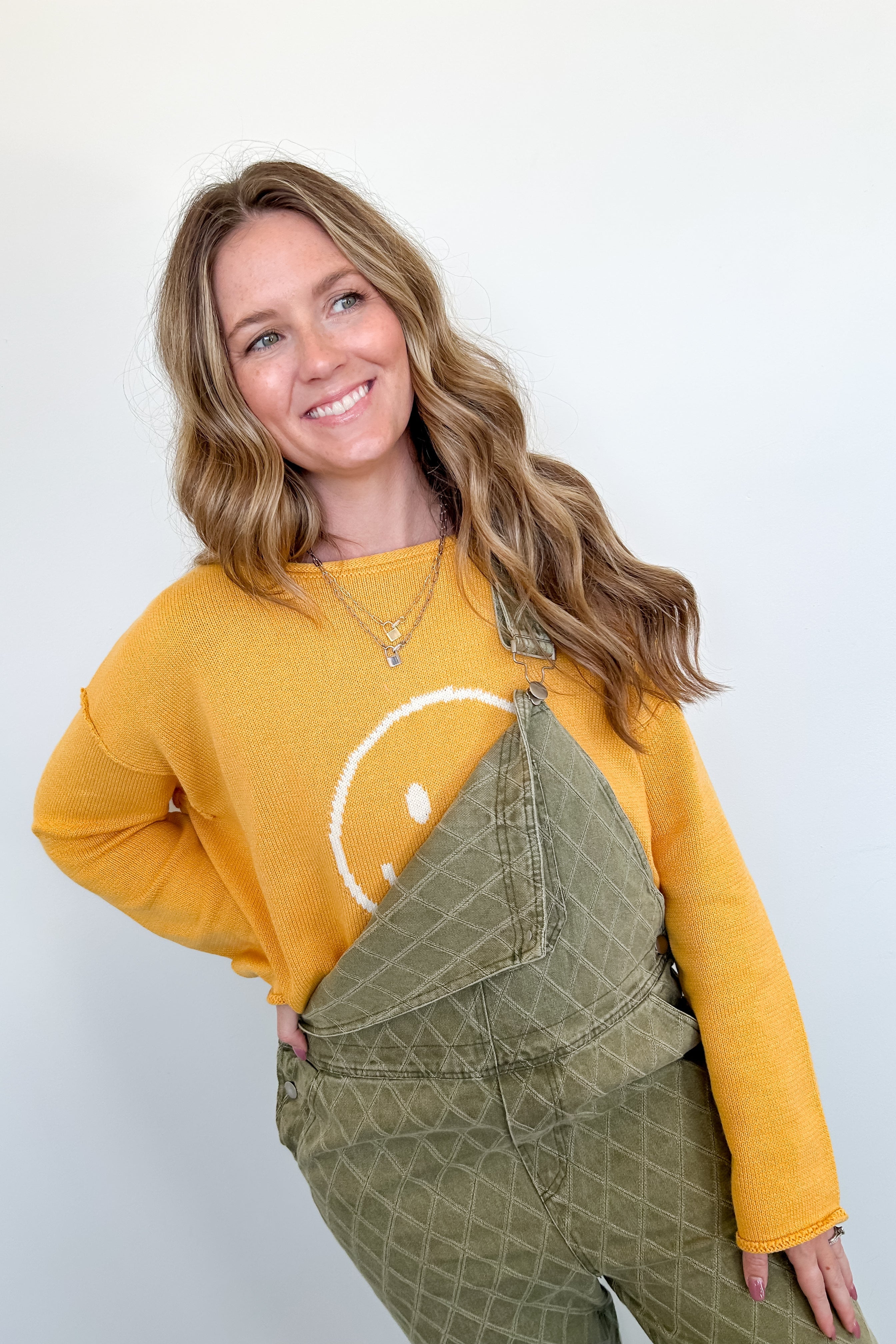 Woman wearing a yellow sweater with a smiley face and green overalls against a white background