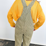 Person wearing a yellow shirt and green patterned overalls standing on a blue mat with a white wall background.