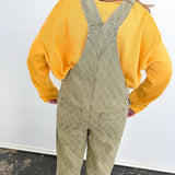 Person wearing a yellow shirt and green patterned overalls standing on a blue mat with a white wall background.