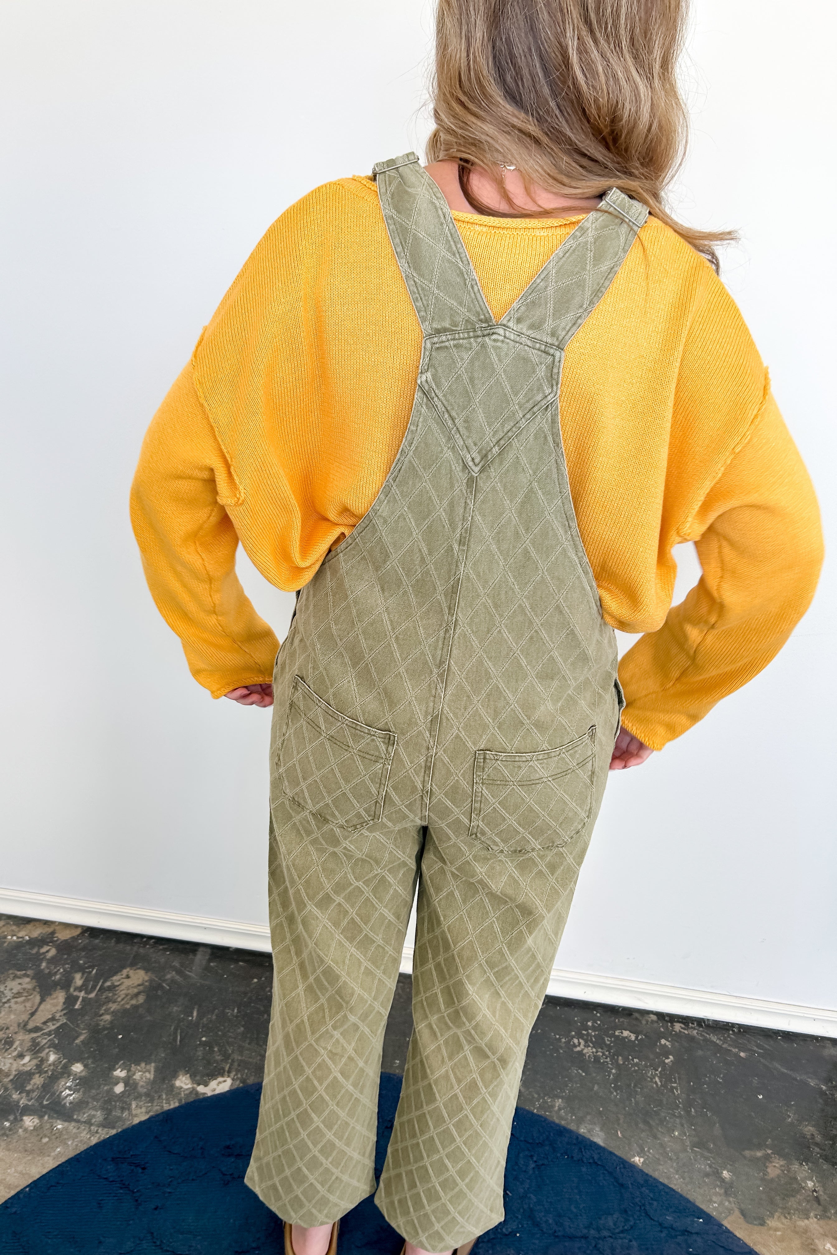 Person wearing a yellow shirt and green patterned overalls standing on a blue mat with a white wall background.