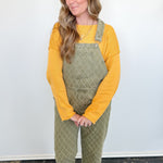 Woman wearing a yellow long-sleeve shirt and green checkered overalls standing against a white wall.