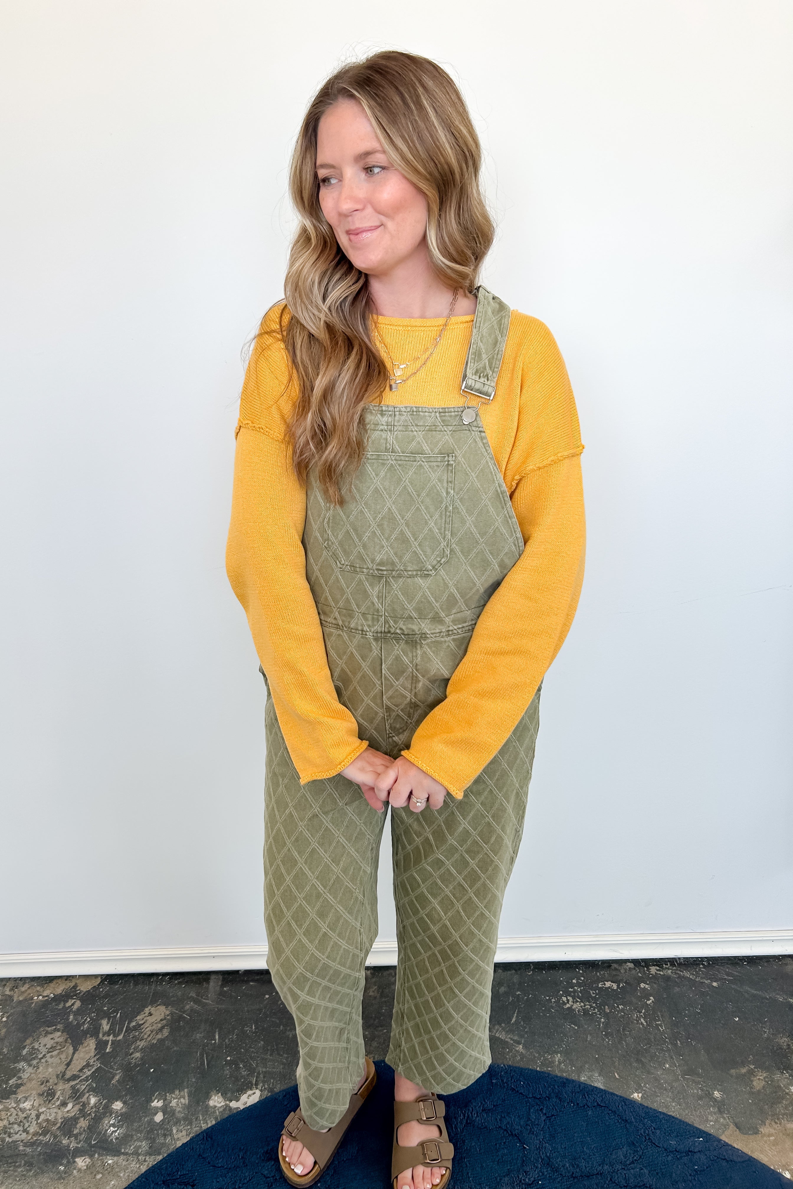 Woman wearing a yellow long-sleeve shirt and green checkered overalls standing against a white wall.